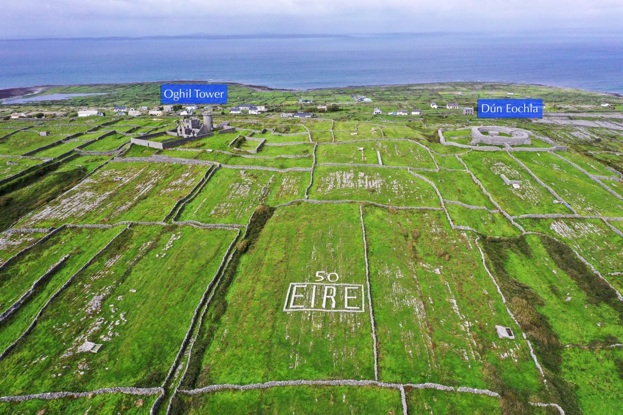 Birds View Ancient Stone Structures among green fields on an island typical Irish costal scenery with overlay blue labels Dun Eochla on the right and Oghil Tower on the left stone walls create lots of small fields in the middle Eire 50 sign made on one of the fields to commemorate 50 anniversary of Ireland's independence 